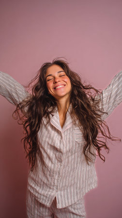 A young woman stands with her arms wide open, smiling happily in soft pajamas. The pastel pink background adds a cheerful vibe, perfect for a relaxed morning atmosphere..の素材