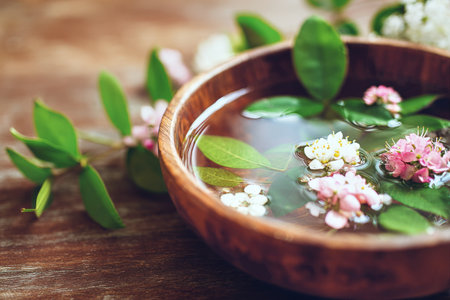 A wooden bowl holds delicate white flower petals and green leaves floating in still water, illuminated by soft light that creates a peaceful atmosphere..の素材