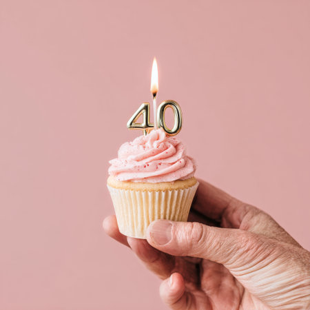 A hand holds a delicious pink frosted cupcake topped with a golden candle reading 40. The soft pink background adds to the festive atmosphere of a birthday celebration.の素材