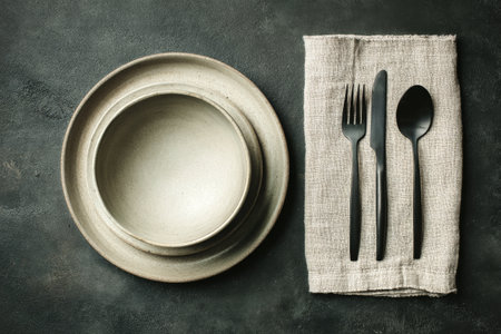 Three ceramic plates of varying sizes are stacked neatly on a gray cloth. A knife and fork with a sleek black finish are placed beside the plates, creating a stylish dining arrangement..の素材