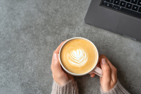 Sipping a beautifully crafted latte with heart-shaped art while sitting at a table with a laptop nearby, perfect for a relaxed work session.の素材