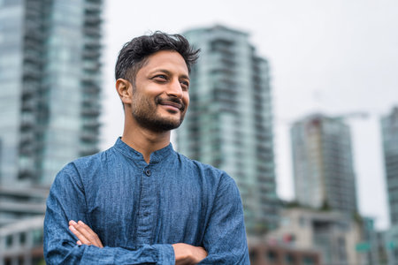 A young man stands confidently with his arms crossed in front of contemporary architecture. The weather is cloudy, creating a calm atmosphere in the urban setting..の素材
