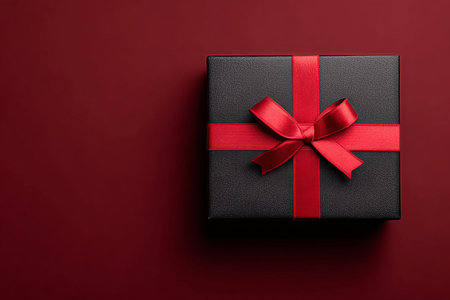 A neatly wrapped black gift box tied with a bright red ribbon sits on a deep red surface, perfect for celebrations or special events. The elegant design highlights joy and anticipation.の素材