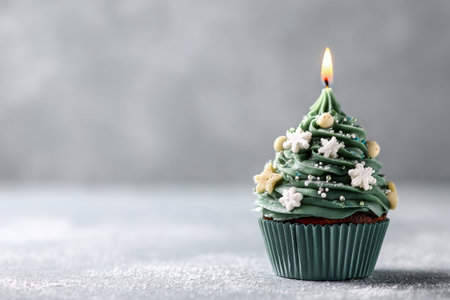 A delightful cupcake designed to look like a Christmas tree sits on a light gray surface. Snowflakes adorn the green frosting, with a candle glowing softly nearby..の素材