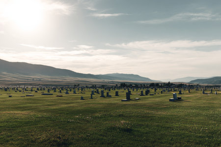 A wide view of a cemetery filled with headstones, set against a backdrop of rolling hills and mountains. The sun casts a gentle light on the green grass.の素材