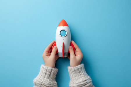 A child with hands gripping a bright toy rocket featuring orange and white colors. The rocket is set against a vibrant light blue surface, creating a fun scene..の素材