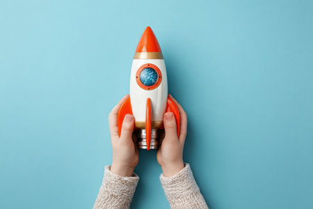 A child with hands gripping a bright toy rocket featuring orange and white colors. The rocket is set against a vibrant light blue surface, creating a fun scene.の素材