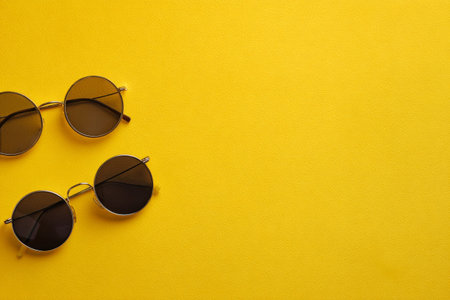 Two pairs of black round sunglasses are placed on a bright yellow surface. The simple arrangement emphasizes the stylish design and bold colors, creating an eye-catching display..の素材