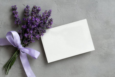 A small bouquet of lavender tied with a light purple ribbon sits beside a blank card on a smooth gray surface, perfect for writing notes or messages for various occasions.の素材