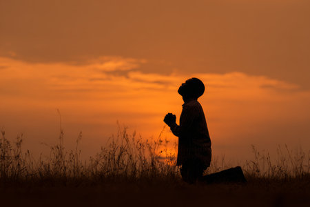A man kneels in quiet prayer on grass, silhouetted against a vibrant sunset. The sky glows with hues of orange and gold, creating a serene atmosphere.の素材