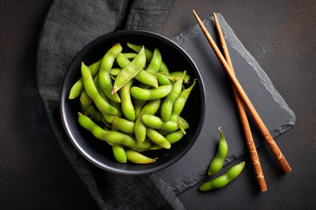 A bowl filled with vibrant green edamame pods is placed on a textured surface. Wooden chopsticks lay beside the bowl, enhancing the presentation of this healthy snack..の素材