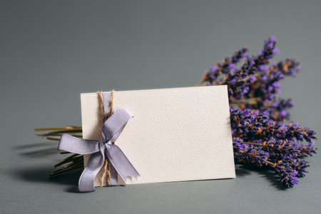 A small bouquet of lavender tied with a light purple ribbon sits beside a blank card on a smooth gray surface, perfect for writing notes or messages for various occasions..の素材