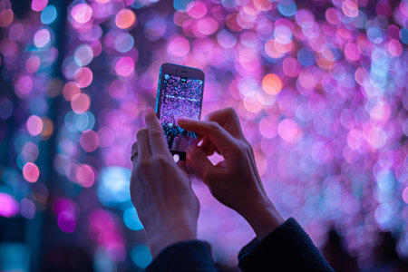 A person holds a smartphone, capturing bright bokeh lights in a city at night. The colorful lights create a festive atmosphere, filling the surroundings with joy.の素材