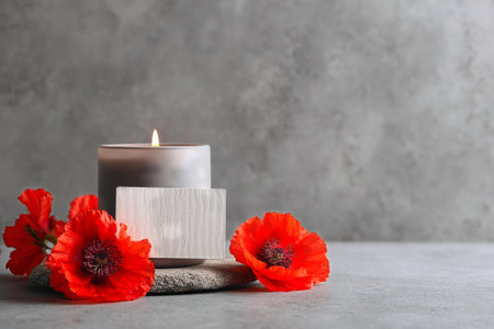 A lit candle sits in a glass holder surrounded by vibrant red poppy flowers. The warm glow combined with floral beauty enhances a calm and inviting ambiance..の素材