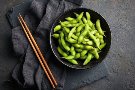 Bright green edamame pods fill a black bowl, set against a dark stone surface. Two wooden chopsticks lie nearby, perfect for enjoying this healthy snack at any time..の素材