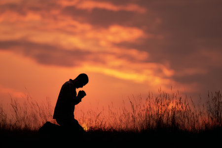 A man kneels in prayer against a vibrant sunset sky, surrounded by tall grass. The serene atmosphere evokes a sense of peace and reflection at dusk..の素材
