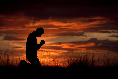 A man kneels in prayer against a vibrant sunset sky, surrounded by tall grass. The serene atmosphere evokes a sense of peace and reflection at dusk.の素材