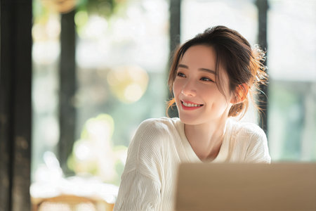 A cheerful woman with brown hair sits at a table in a modern cafe, focused on her laptop. Sunlight streams through large windows, creating a warm atmosphere..の素材