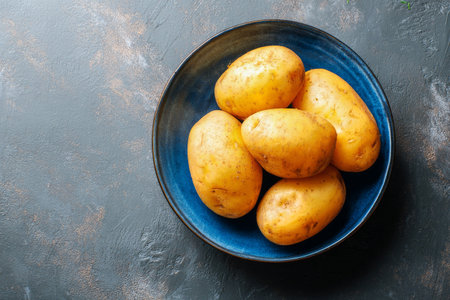 A bowl filled with freshly harvested yellow potatoes sits on a dark textured surface, showing their natural skin and earthy appeal. Perfect for cooking and nutrition.の素材
