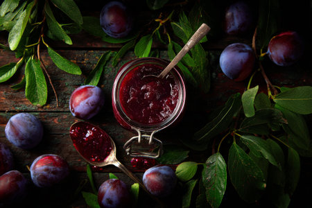 A jar of rich plum jam sits on a wooden board, surrounded by juicy ripe plums and green leaves, highlighting the colors and textures of the ingredients in a natural setting..の素材