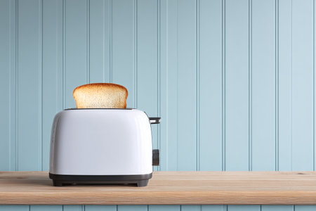A white toaster is sitting on a kitchen counter with a slice of golden brown toast popping up. The walls are painted light blue, creating a bright and inviting atmosphere..の素材