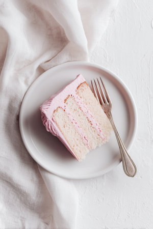 A slice of fluffy cake with pale pink frosting sits on a round plate. A silver fork is next to the cake, inviting dessert lovers to enjoy a sweet treat.の素材