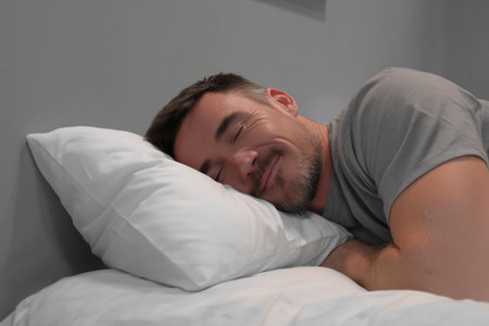A man is lying on his side, smiling while resting his head on a soft pillow. The room features neutral colors and a serene atmosphere, ideal for relaxation.の素材