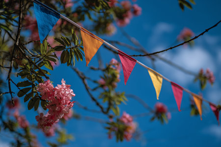 Colorful triangular bunting hangs between branches of a flowering tree. The scene captures the vibrancy of spring, with bright flags contrasting against blue sky and blossoms.の素材