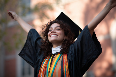 A young woman in a graduation gown and a colorful sash smiles broadly as she celebrates her achievement. The ceremony takes place outdoors under a clear blue sky.の素材