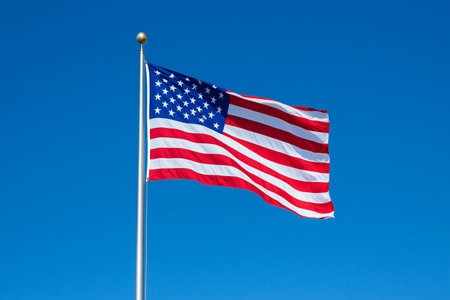 The American flag flutters gently on a flagpole under a bright blue sky. The vibrant colors of the flag stand out beautifully against the clear backdrop of a sunny day.の素材