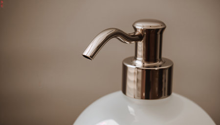 A pump bottle sits on a bathroom counter. It has a shiny metal pump and a soft colored bottle. The background shows a tiled surface. Light reflects off the pump.の素材