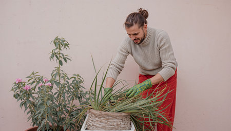 A man is working with potted plants in a garden. He is wearing gloves and is focused on caring for the greenery. It is daytime, and a bush is in the background.の素材