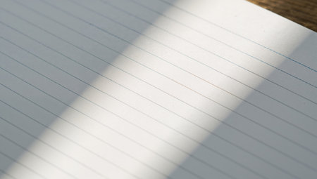 Sunlight shines through a window, creating a shadow on a sheet of lined paper laying flat on a wooden table. The scene shows a simple indoor setting during daytime.の素材