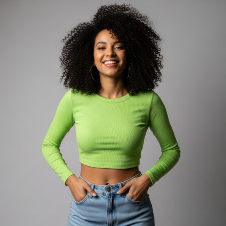 A studio portrait captures a young, beautiful woman with dark, voluminous curly hair. She stands facing forward, smiling radiantly directly at the camera, exuding happiness and confidence. Her attire consists of a bright lime green, form-fitting, long-sleeved crop top paired with light blue denim jeans. She has large gold hoop earrings. Her hands are casually placed in the front pockets of her jeans. The background is a simple, neutral gray, which highlights her bright outfit and smile. The lighting is soft and even, typical of a professional studio shot.の素材