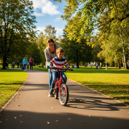 A mother is seen helping her young child ride a bicycle in a serene park setting. The child is seated on the bike, wearing a helmet, while the mother stands behind, offering support and guidance. The path is surrounded by lush green trees and grass, with other park-goers visible in the background. The scene captures a moment of learning, bonding, and outdoor enjoyment.の素材