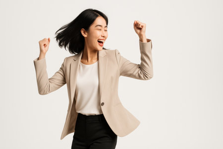 Side profile to three quarter turn happy young Asian business woman in a beige blazer spinning with both hands raised celebratory studio portrait on whiteの素材
