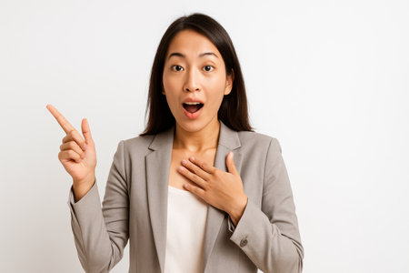 Asian businesswoman threequarter right angle hand on chest and raised hand reacting to big news amazed expression facing camera studio portrait isolated on whiteの素材