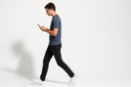 Young man using a smartphone side profile mid step with the front foot lifted and the phone held at chest height on whiteの素材