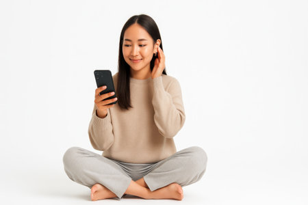 Young Asian woman in cozy loungewear sitting cross legged on white floor with smartphone on clean background relaxed home lifestyle concept for social media streaming and digital wellbeingの素材