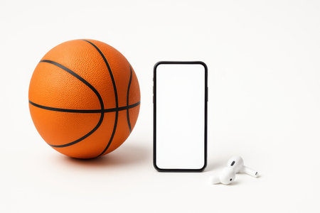 Studio photo orange basketball ball placed beside modern smartphone with blank white screen and simple wireless earphonesの素材