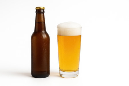 Unlabeled brown beer bottle standing beside clear glass of golden beer with frothy foam on glossy white backgroundの素材