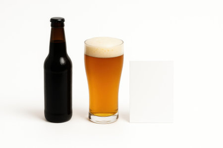 Unlabeled dark beer bottle placed behind tall clear glass of beer with rich creamy foam on pure white backgroundの素材