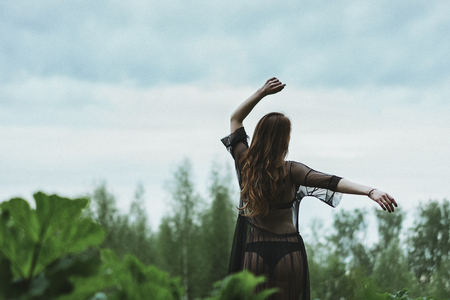 A dark-haired woman in a translucent black dress dancing against the sky and green grass. Relaxation and dreams.の写真素材