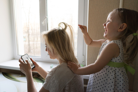 Mom and daughter are sitting by the window. Mother plays in the phone, daughter makes mom hairstyleの写真素材