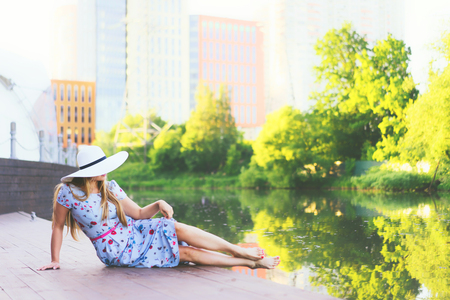 A long-haired woman in a white hat and a beautiful long dress is sitting on the pier by the river in the sity. Concept of summer serene rest. Back viewの写真素材