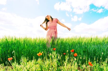 Long-haired woman in a pink dress on a field of green wheat and wild poppies. The concept of freedom and freshness. Beautiful wheat, great design for any purposes. Female symbol. Agriculture harvestの写真素材