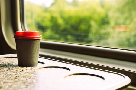 A paper coffee cup with a red plastic lid and a white inscription circle. Coffee to go on a table in the train overlooking a beautiful rural green landscape. Travel, lifestyleの写真素材