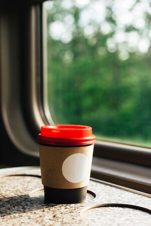 A paper coffee cup with a red plastic lid and a white inscription circle. Coffee to go on a table in the train overlooking a beautiful rural green landscape. Travel, lifestyleの写真素材
