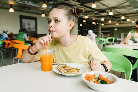 A teenager girl sits in a cafe drinking a carrot smoothie and eating pancakes with a salad. Eating outの写真素材