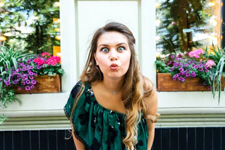 Beautiful young blonde woman in a green summer dress stands against the background of a window of a...